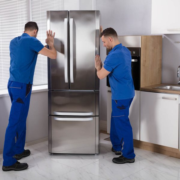 Two Young Male Movers Placing Steel Refrigerator In Kitchen